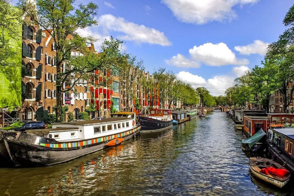 Sunny Amsterdam canal with historic warehouses, red shutters, and houseboats under blue sky with clouds